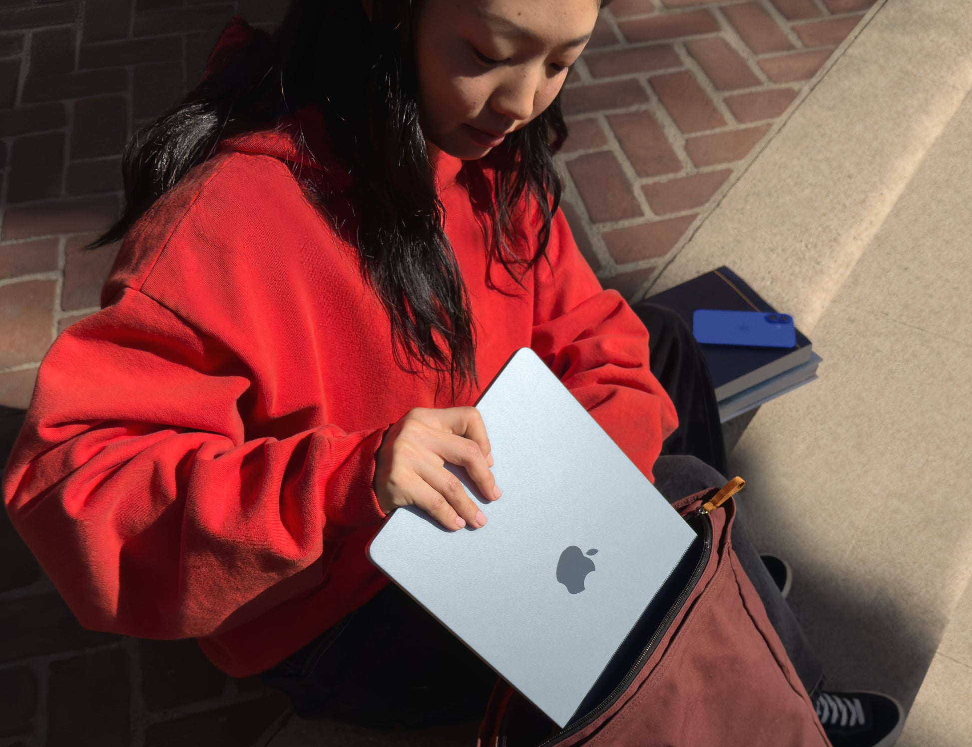 Person placing a 13 inch MacBook Air Midnight into a backpack while sitting outdoors on brick pavement